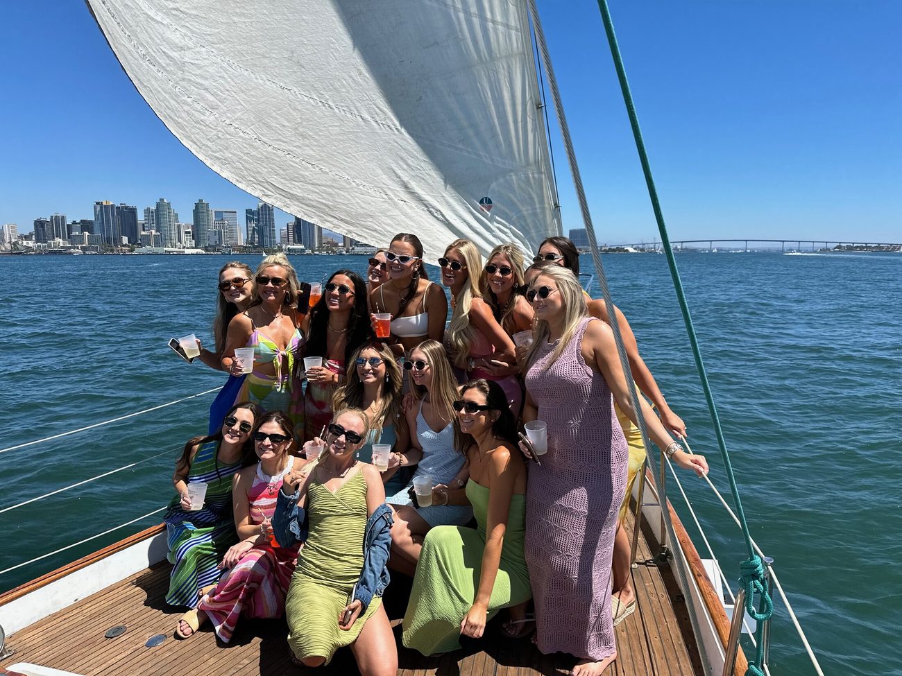 A bachelorette group in bright summer dresses on JADA's bow, sails up, the San Diego skyline behind them